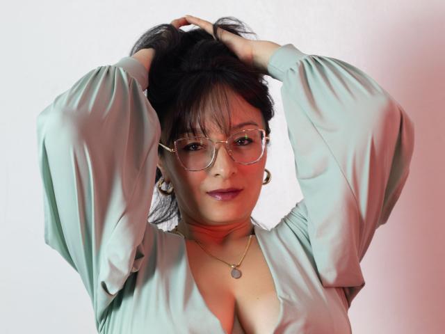 LexiLuna with hands in black hair, wearing glasses, green blouse, and gold hoop earrings, showing cleavage, smiling. Soft light, red highlight on cheek. Cute and sexy.