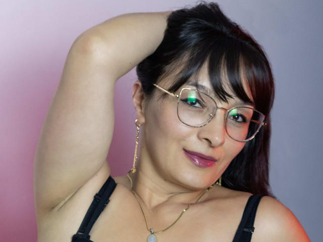 LexiLuna with glasses, dark hair, and pink lipstick, raising her arm, showing her armpit, wearing a black strap top, and gold earrings, smiling slightly.