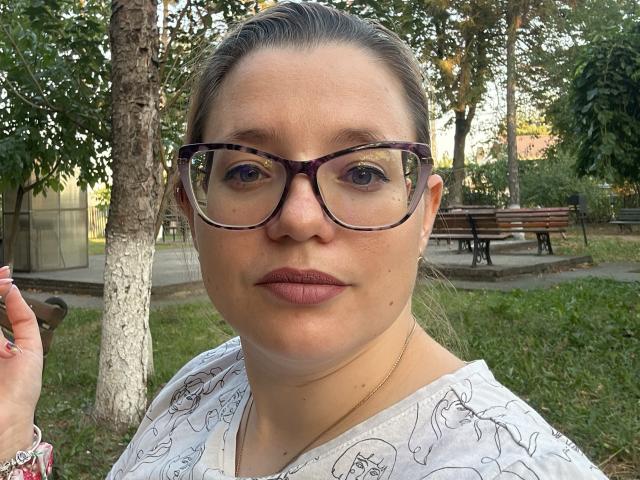 MiaBlue69 with glasses, white patterned shirt, brown hair in a bun, pinkish lipstick, and a gold necklace, standing in a park, looking at the camera with a neutral expression.