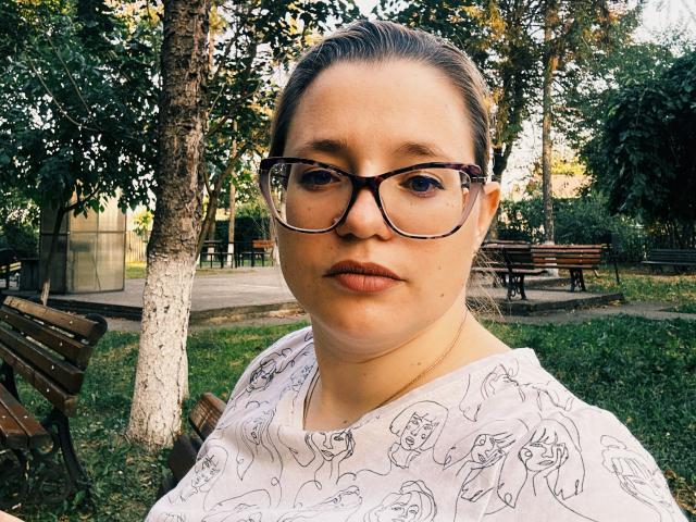 MiaBlue69 with glasses, pale skin, and pouty lips, wearing a white patterned shirt, standing in a park, has a chubby face and brown hair pulled back.