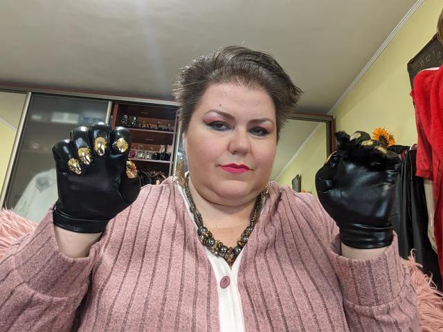 KaryQueen with short brown hair, pink cardigan, white shirt, black leather gloves, gold-accented nails, pink lipstick, and a necklace, holding gloves up. Her face is slightly chubby.