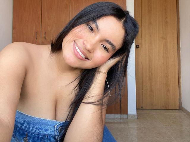 MiiaPalmer with dark straight hair, smiling, head resting on hand, wearing denim top, showing her boobs, brown eyes, tan skin, gold bracelet, wooden doors in background.
