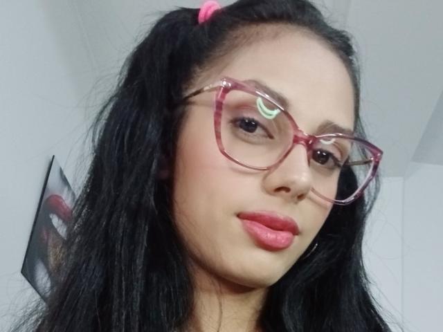 AnaisHilton with long black hair in pigtails, wearing pink glasses and pink lipstick, has a cute face and is looking slightly to the side with a neutral expression.