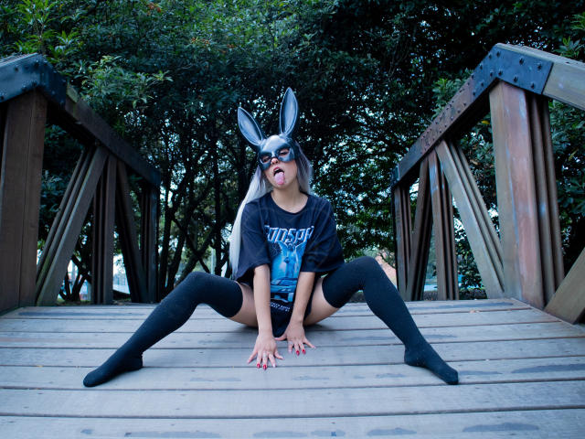 MaveParker with gray bunny mask, black shirt, blue graphic, black thigh-highs, and bare ass, sitting on wooden bridge, legs spread, tongue out, surrounded by green trees.
