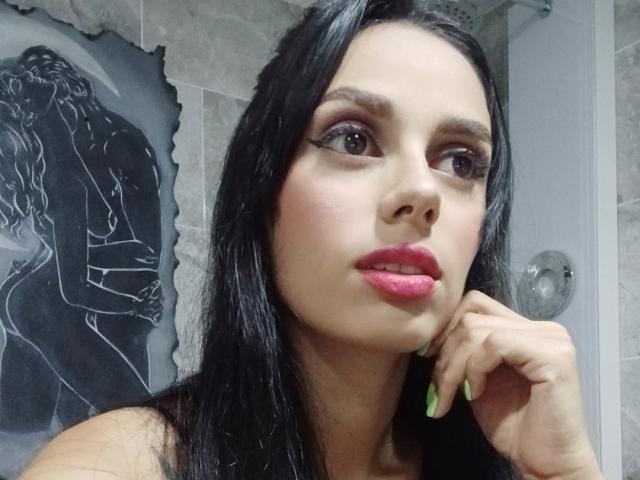AnaisHilton with black hair, pink lipstick, green nails, and dark eye makeup, looking at camera with hand on cheek, in a bathroom. Gray tiled walls, black-and-white wall art.