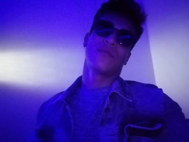Lestatd with spiked dark hair, wearing sunglasses, a grey shirt, and a denim jacket, under purple blue lighting. Lestatd has a slight smirk and a nose ring.