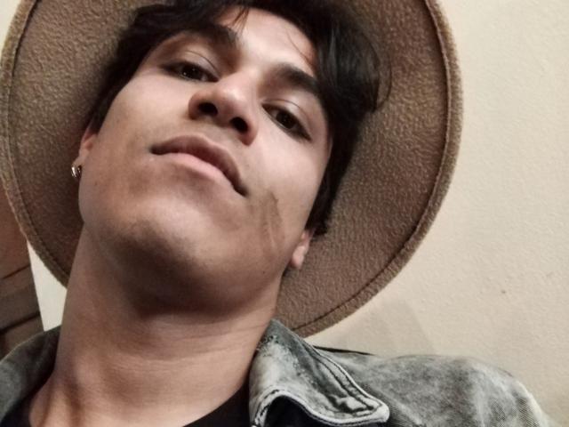 Lestatd with a brown hat, wearing a gray jacket, has dark hair and a small earring in his left ear, looking up with a slight smile.