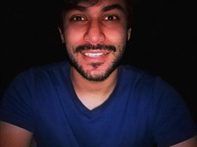 Lestatd has a dark, curly mustache, a slight beard, and is wearing a blue V-neck shirt. He's smiling with his teeth showing, against a black background.