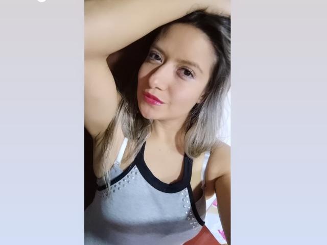 ValeriaHunter69 with grey and black sports top, pink lipstick, arm raised, grey-brown hair, slight smile, visible nipples, light skin, medium breasts, soft lighting, casual selfie.