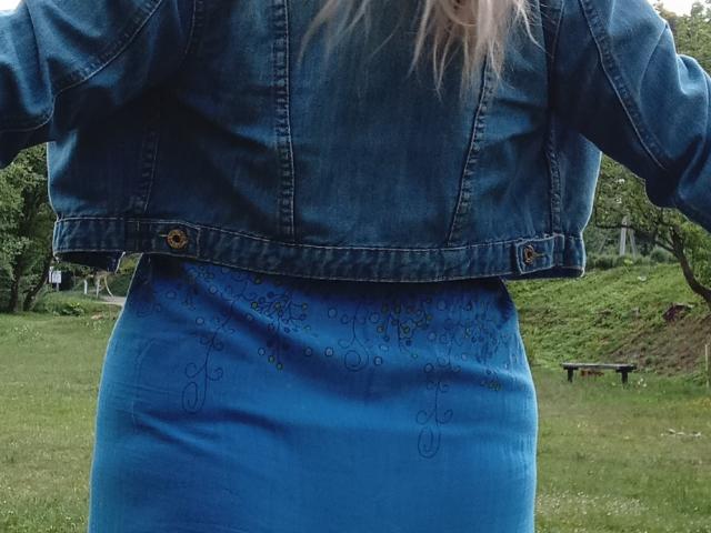 KelliRedhaers blaue Jeansjacke und der dazu passende blaue Rock, der ihr Gesäß zeigt, mit dezenten Stickereien auf dem Rock. Ihr blondes Haar ist teilweise sichtbar..