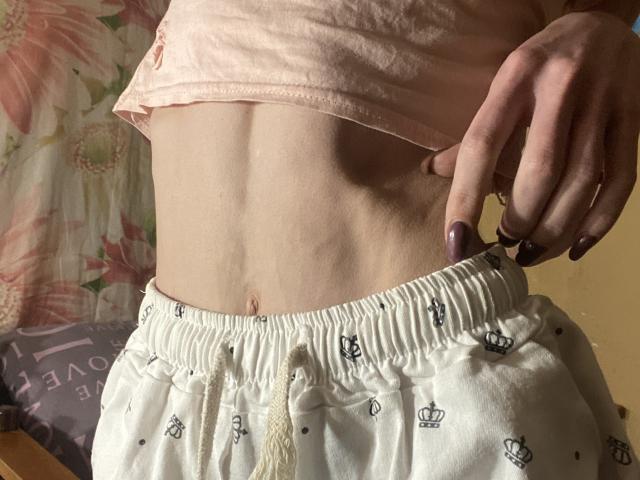 Kabblasha's flat stomach, wearing white pants with crown prints, lifting a pink shirt, showing a small navel, with purple nail polish on her right hand.