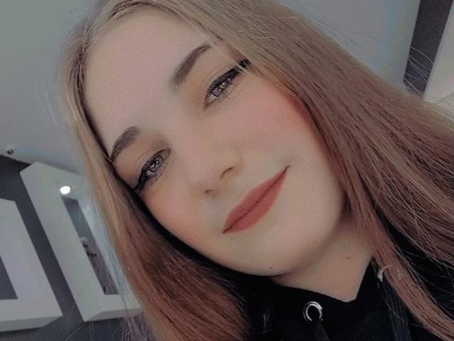 NikaStark has long blonde hair, light skin, and is smiling with subtle makeup, including black eyeliner and pink lipstick. She's wearing a black shirt with a button.