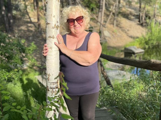 HelenAndChristy, curly blonde, wearing red sunglasses, purple tank top, black pants, standing by white birch, holding it with her right hand, smiling, outdoors, sunny, wood path.
