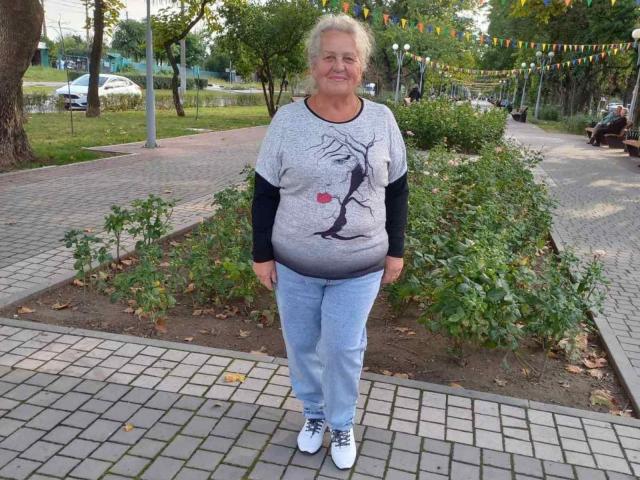 HelenAndChristy has a chubby belly, wears a grey tee with a black tree and red heart, blue jeans, and white sneakers, standing on a brick pathway.