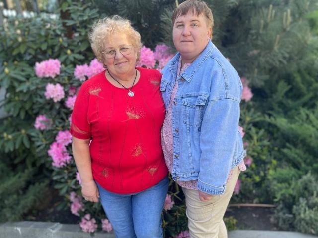 HelenAndChristy with curly blonde hair, glasses, red shirt, and blue jeans stands next to a chubby, brown-haired guy in a blue denim jacket and pink shirt.