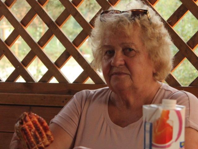 HelenAndChristy with curly white hair, wearing a pink shirt, holding a waffle, and a pink and white drink in front of a wooden lattice. Sunglasses on head.