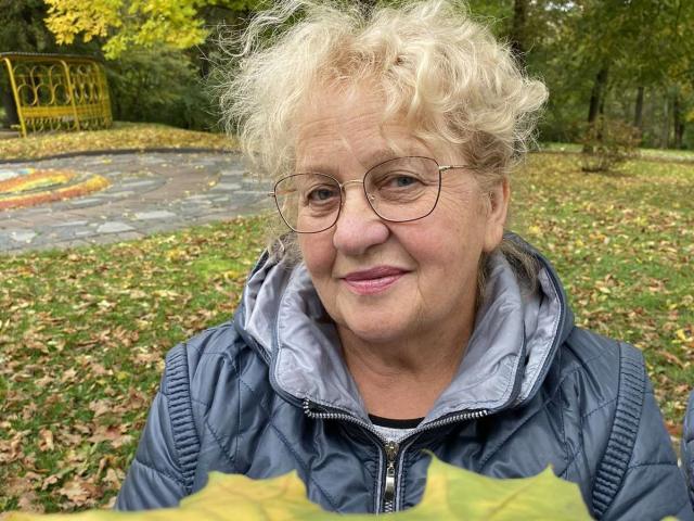 HelenAndChristy with curly blonde hair, glasses, and pink lipstick, wearing a blue puffy jacket, holding yellow leaves, smiling in a park.