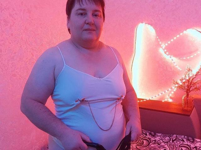 HelenAndChristy has a chubby, pale body with small tits, wearing a white camisole with metal nipple clamps and a chain, holding black underwear, in pink and purple light.