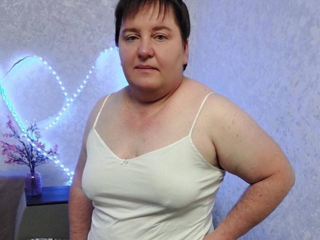 HelenAndChristy, a chubby white woman with short brown hair, wearing a white spaghetti-strap top, has her hands on her hips, showing her tits and a confident expression.