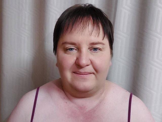 HelenAndChristy has short dark hair, light skin, small blue eyes, and a slight smile. She's wearing a pinkish top with thin purple straps, showing her tits.