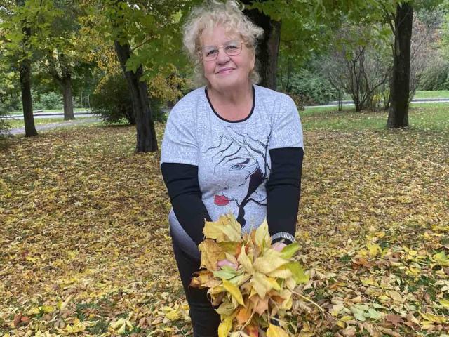 HelenAndChristy has curly blonde hair, glasses, and a grey shirt with a black graphic, holding a pile of yellow and brown autumn leaves. She's smiling.
