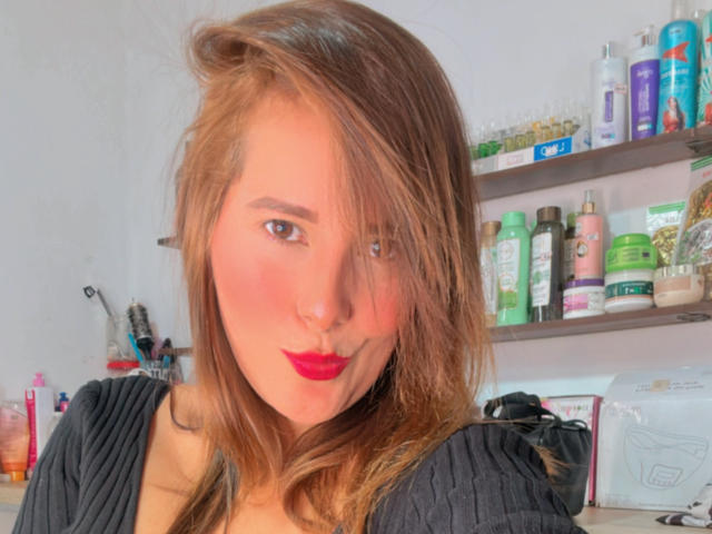 SaritaVelez has brown hair, red lipstick, and a pouty face with a nose ring, wearing a black top, with makeup and toiletry items in the background.