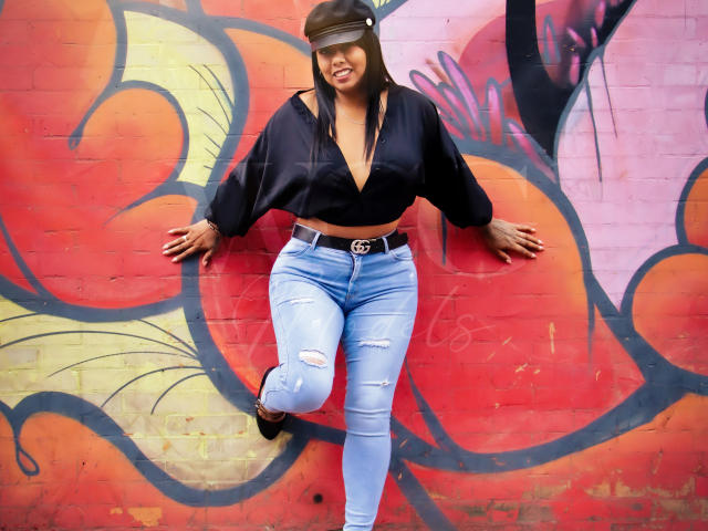 LizHerrera has big tits, wearing a black blindfold, black crop top, and ripped blue jeans with a Gucci belt, standing against a colorful graffiti wall.