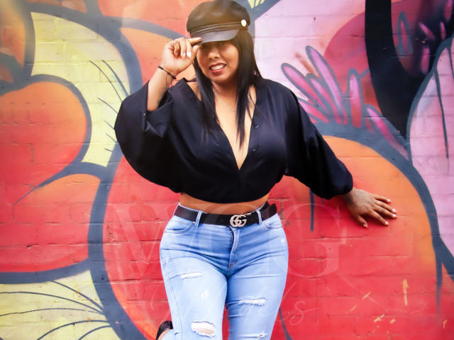 LizHerrera has long black hair, wearing a black cap, deep V-neck blouse, and ripped blue jeans with a Gucci belt. She's smiling, adjusting her cap, with medium tits.
