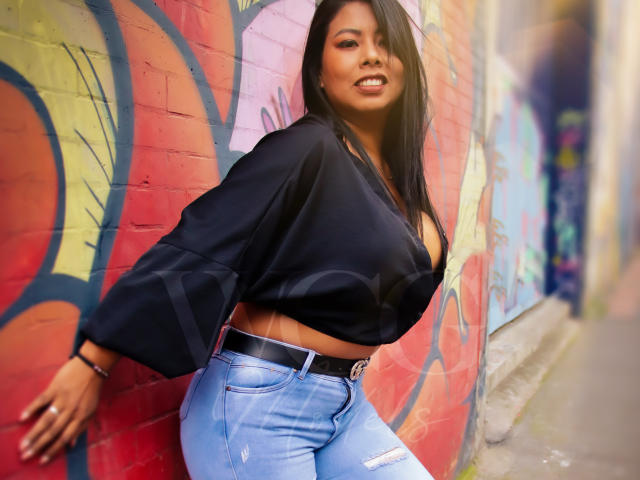 LizHerrera with medium tits, wearing a black crop top and blue ripped jeans, leaning against a graffiti-covered wall, smiling. She has long black hair and a black belt.