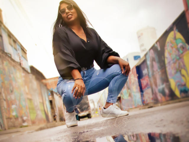 LizHerrera with dark skin, long black hair, wearing sunglasses, black blouse, blue ripped jeans, and white sneakers, squatting in a graffiti-laden alleyway with a reflective puddle.