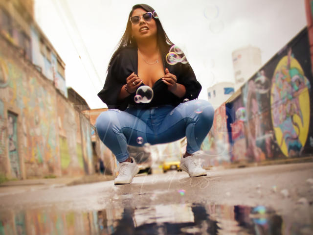 LizHerrera squats on a wet street, wearing a black top with her tits exposed, blue jeans, and white sneakers, blowing bubbles with her hands. She's got long dark hair.
