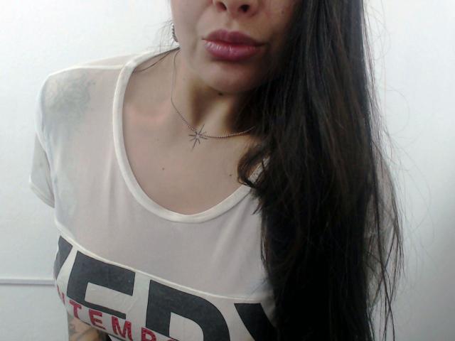 Gemma Lopez - aantrekkelijk, met lang donker haar, roze lippen, een wit, doorschijnend topje dat haar borsten laat zien, zwarte letters en een spinnenketting. Tattoo op de linker schouder. Witte muur als achtergrond..