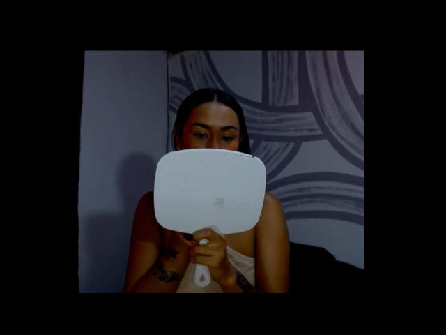 DaphneJill tiene in mano un telecomando bianco e rettangolare davanti a sé, coprendosi il seno, con i capelli castani sciolti e tatuaggi visibili sul petto..