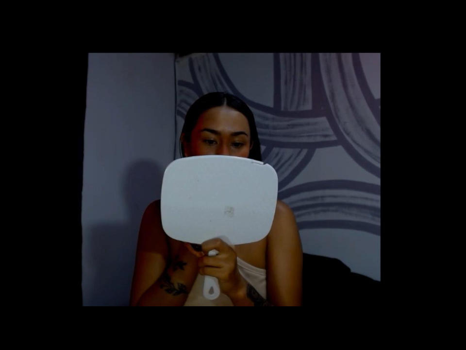 DaphneJill hält eine weiße, rechteckige Fernbedienung vor sich, die ihre Brüste bedeckt, mit offenem, braunem Haar und sichtbaren Tattoos auf ihrer Brust..