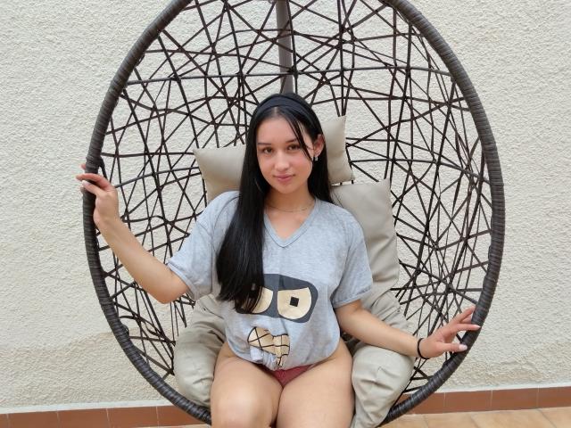ValeriiNaughty with long black hair, wearing a gray tee with a cartoon face and red panties, sitting in a black hanging chair with a white cushion.