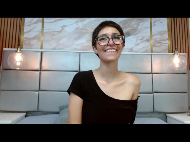 MaiGolf with glasses, smiling, black off-shoulder top, small tits, short dark hair, sitting in front of a bed with two hanging lamps.