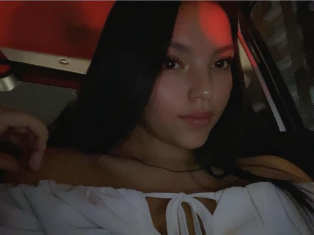IssaPuasini has long black hair, medium brown skin, and wears a white off-shoulder top with a visible chest. Red light casts a warm glow on her face.