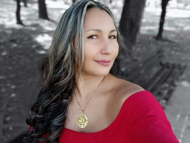 ShaaronCool with long black-and-gray hair, wearing a red off-shoulder top, gold necklace with circular pendant, smiling at camera, showing subtle makeup and pink lipstick.
