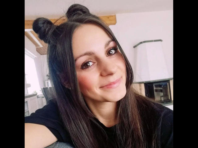 LoellaRose-hot has dark hair in two buns, brown eyes, pink blush, and a cute smile. She's wearing a black shirt, looking at the camera, in a bright room.