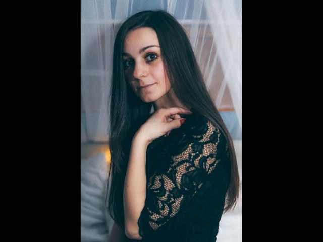 LoellaRose-hot with long brown hair, wearing a black lace top, has her hand near her chin, smiling softly. She has light skin and brown eyes, looking cute.