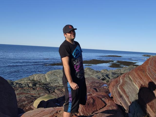 MikeWazowski69 está em uma praia rochosa, vestindo uma camisa preta com um desenho azul e roxo, shorts pretos e um boné preto. No fundo, há um oceano azul e um céu claro. MikeWazowski69 tem um traseiro pequeno..