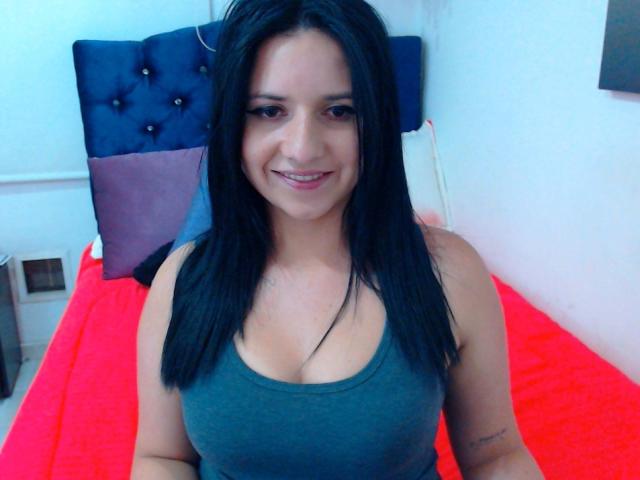 Kkatalina with black hair, green tank top, smiling, showing her boobs, sitting on a red bed, blue and purple headboard, white wall, tattoo on right arm.