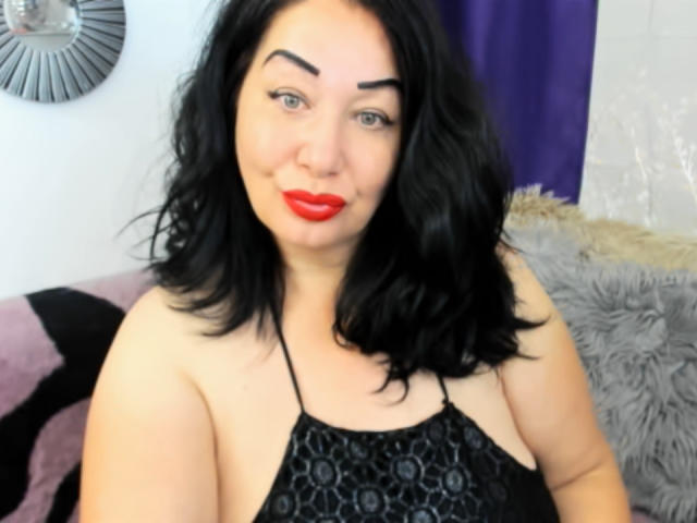 LexyRose a du rouge à lèvres, des cheveux noirs et des sourcils épais. Elle porte un haut noir à encolure dégagée avec un motif, qui laisse apparaître ses épaules nues et une partie de sa poitrine..