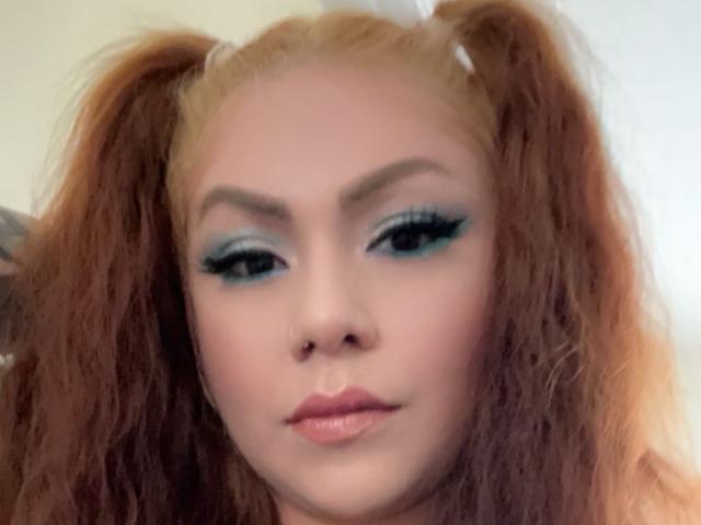 Blush69 hat rote, lockige Haare zu Zöpfen gebunden, trägt blauen Eyeliner und pinke Lippen und blickt direkt in die Kamera mit einem neutralen Gesichtsausdruck. Ihre Nase ist klein und ihre Haut ist hell..