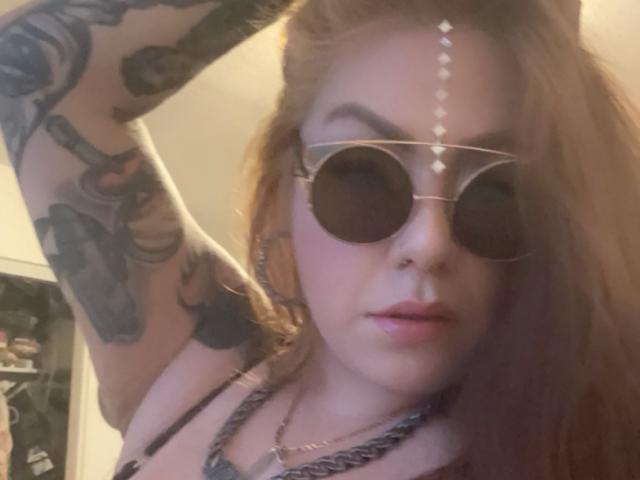 Blush69, avec des tatouages sur son bras, portant des lunettes de soleil rondes et un collier en chaîne, a les cheveux roux et un anneau au nez. Elle lève son bras, montrant sa poitrine et un maquillage discret..