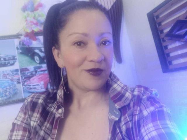 CharloteEzra has dark brown hair in a ponytail, wears a plaid shirt, and has dark lipstick. She's smiling with hoop earrings and a slight blue light.