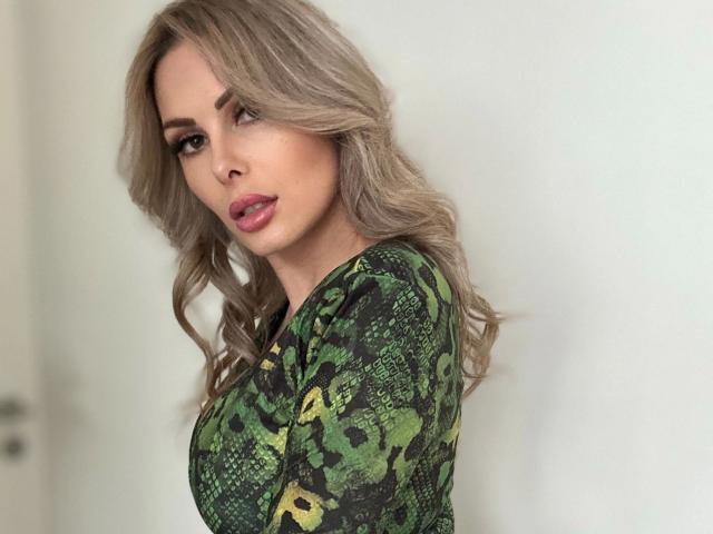 XANIA-WET ha i capelli biondi mossi, una maglietta verde con stampa a serpente e labbra rosa carnose. Ha un seno di taglia media ed è rivolta di spalle con un'espressione seducente..