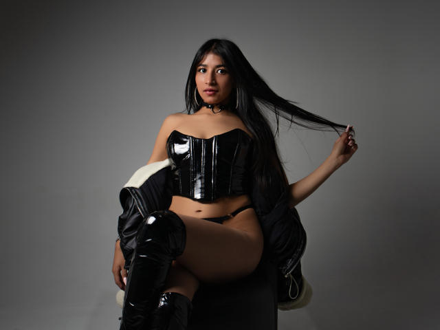NaiaSanders with long black hair, wearing a shiny black leather corset and thigh-high boots, sitting on a chair, holding her hair, showing her ass in black panties.
