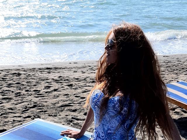 SuperLongLegs69 with long brown hair, wearing a blue patterned top, sitting on a blue-striped chair, facing the ocean, wearing sunglasses, hands on her lap, sunny beach.