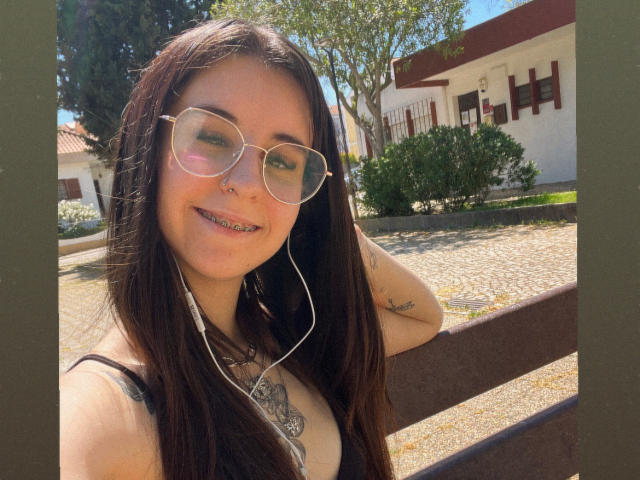 PinkyyFlavour with long brown hair, glasses, nose ring, earbuds, smiling, showing teeth, tattoo on chest, sleeveless top, sunlit, outdoor, brown bench, green trees, white house in background.